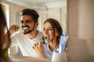 An image of a man and woman brushing their teeth in a bathroom mirror